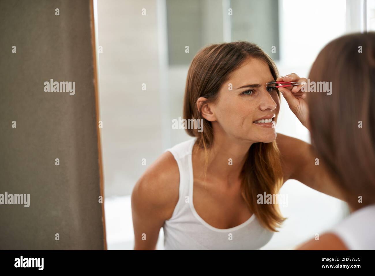 Schön zu sein kann manchmal etwas schmerzhaft sein. Aufnahme einer jungen Frau, die schmerzlich ihre Augenbrauen in einem Badezimmerspiegel zettt. Stockfoto