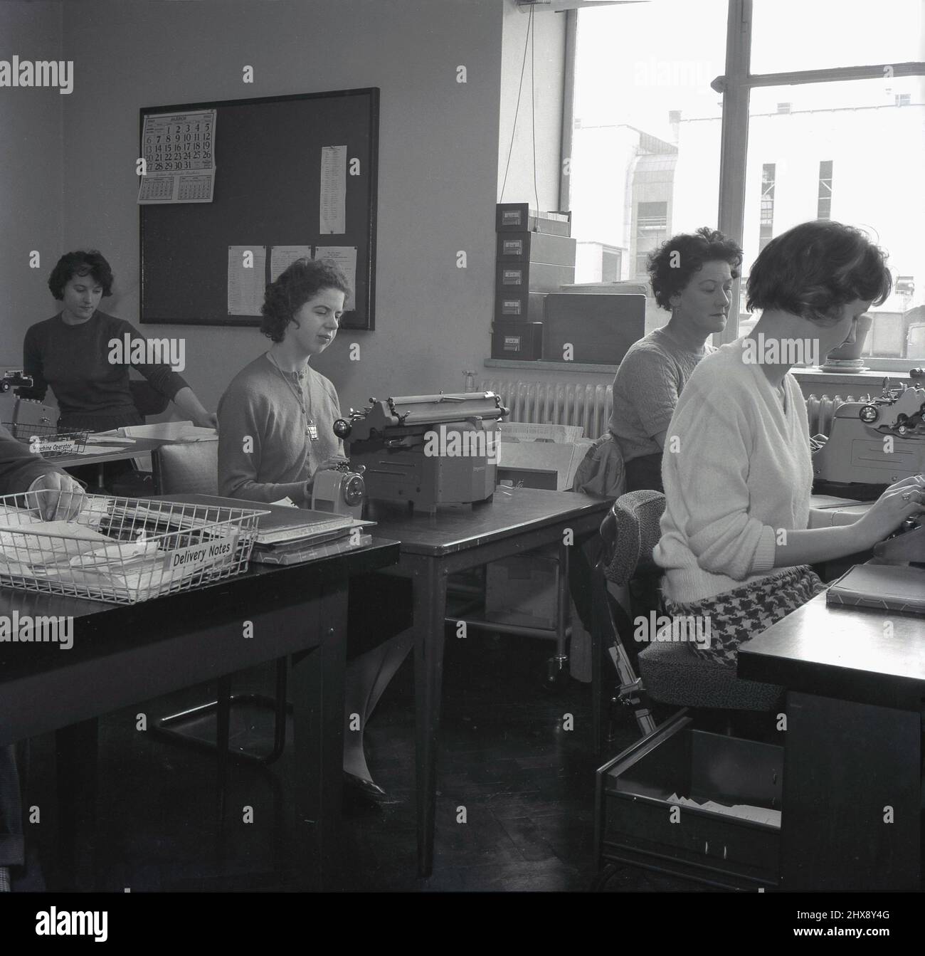 1960, historisch, in einem Verwaltungsbüro in einem Stahlwerk, sitzen vier weibliche Angestellte an Schreibtischen, drei von ihnen mit Schreibmaschinen der damaligen Zeit. Auf einem leeren Schreibtisch, einem Drahteinschub mit einem Etikett mit der Aufschrift „Delivery Notes“, Abbey Works, Port Talbot, Wales, UK. Stockfoto