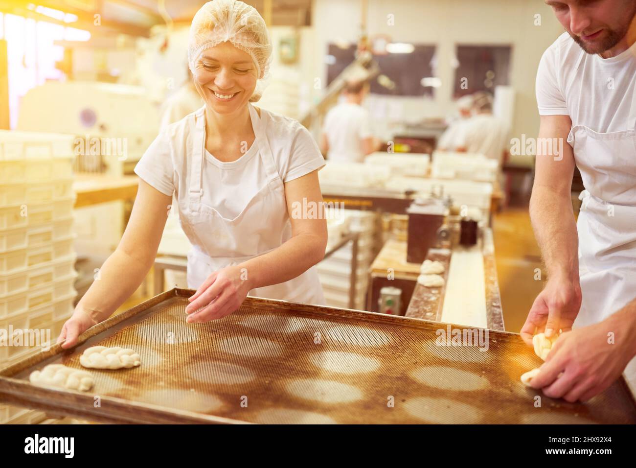 Die junge Frau als Bäckerin der Ausbildung arbeitet in der Bäckerei und legt Hefezöpfe auf ein Backblech Stockfoto