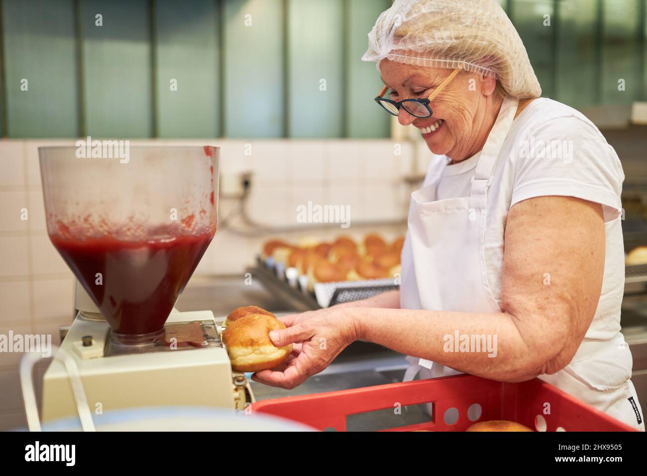 In einer großen Bäckerei füllt eine ältere Frau als Bäckerin Berliner Pfannkuchen mit Marmelade Stockfoto