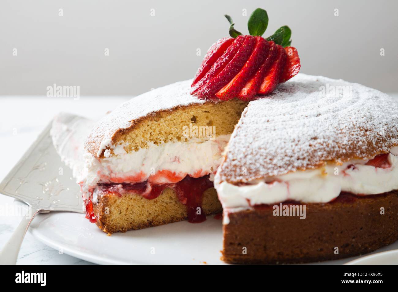 Hausgemachter Viktoria-Biskuitkuchen, gefüllt mit Erdbeeren, Marmelade und Schlagsahne, verziert mit Puderzucker und Erdbeere auf einem weißen Marmortisch Stockfoto