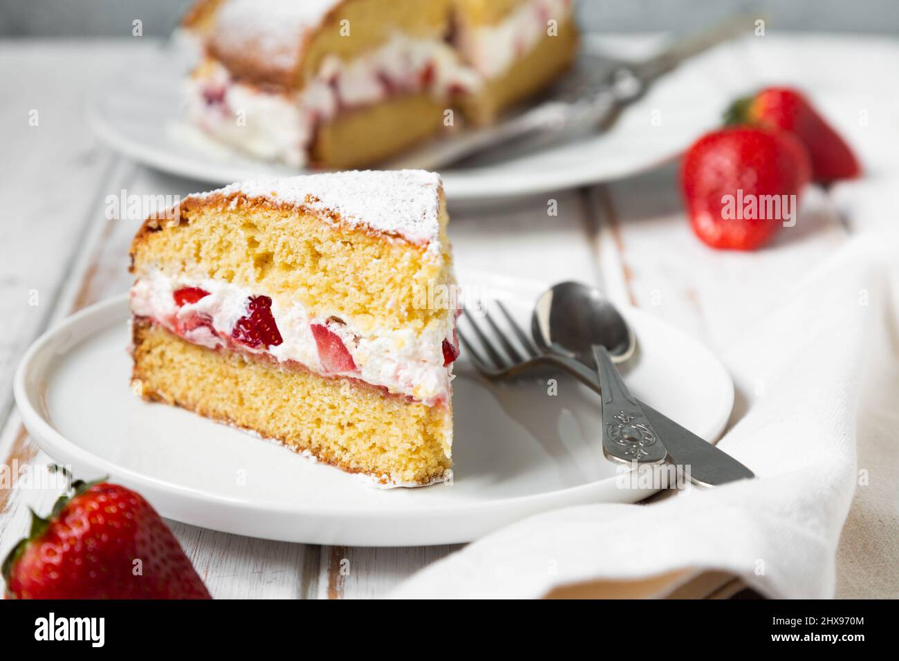 Hausgemachter Viktoria-Biskuitkuchen, gefüllt mit Erdbeeren, Marmelade und Schlagsahne, verziert mit Puderzucker und Erdbeere auf einem weißen Holztisch Stockfoto
