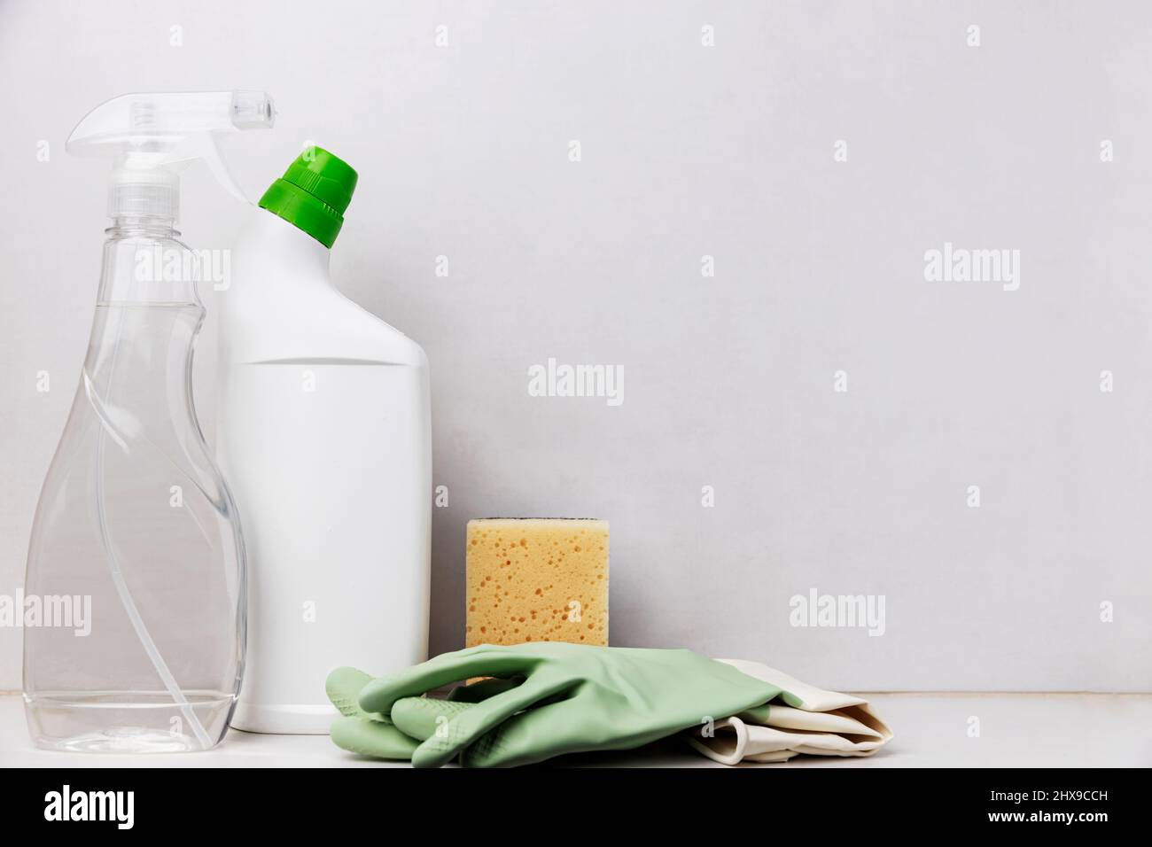 Hausreinigung, Housekeeping-Konzept. Weiße WC-Reinigungsflasche, transparenter Spender-Fensterreiniger, Sanitärware, Waschlappen und grüne Schutzhandschuhe für Hausfrau auf dem Tisch, grauer Hintergrund Stockfoto