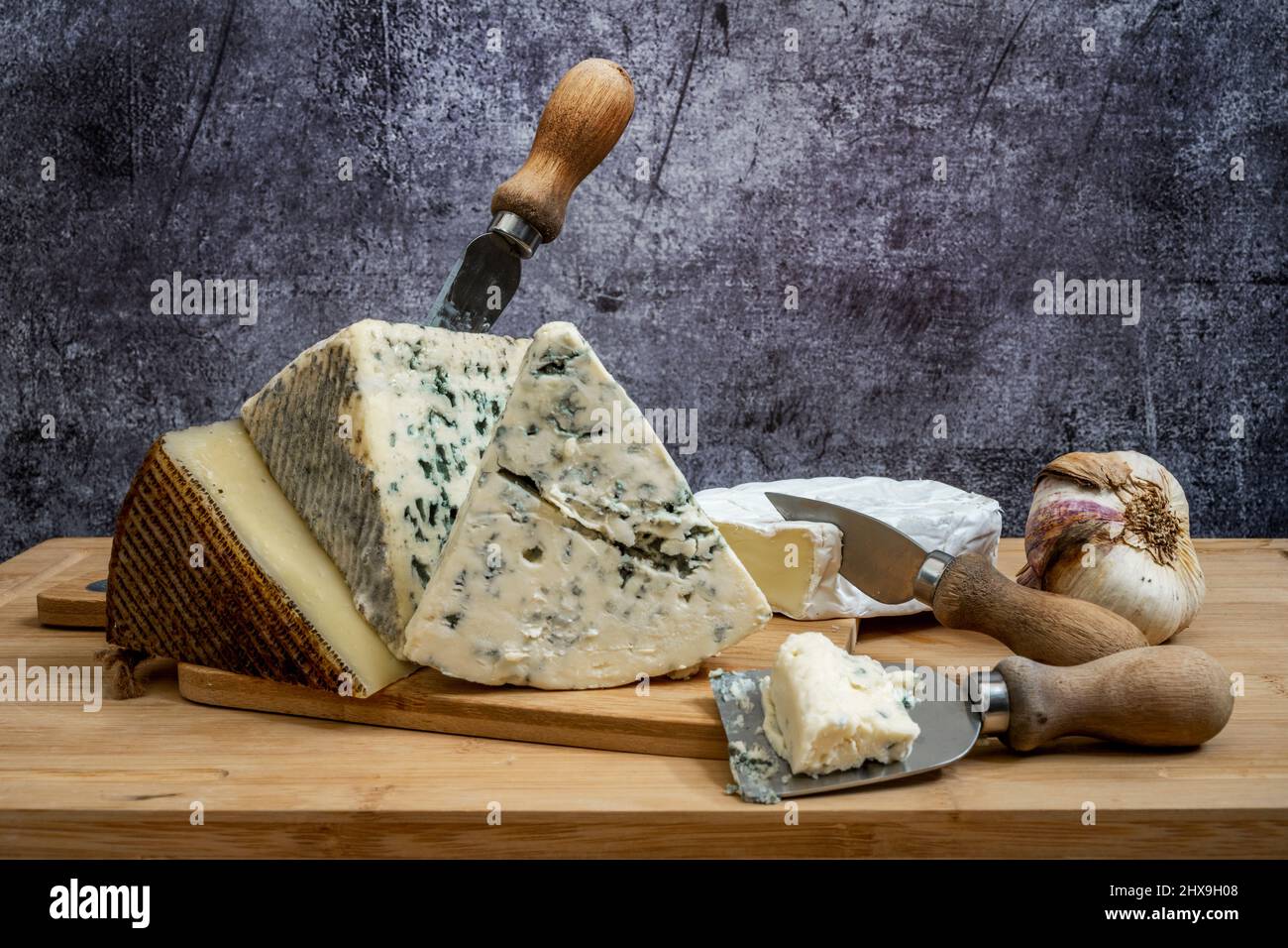 Blauer Kuhkäse auf der Oberseite des ausgehärteten Manchego-Käses, Käsemesser und Knoblauchkopf auf einem Bambusbrett Stockfoto