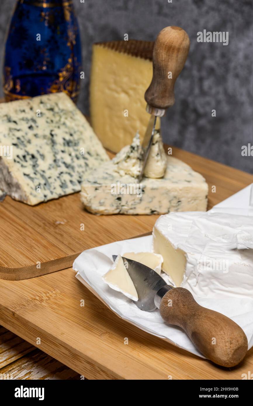 Kleines Käsemesser, das eine Scheibe Brie neben anderen blauen Käsesorten auf einem Schneidebrett aus Bambusholz schneidet Stockfoto