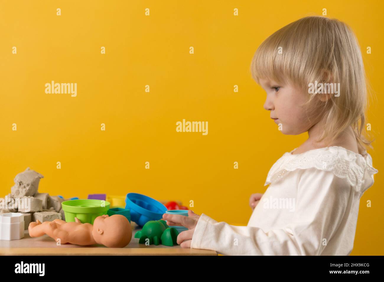 Ein schönes Mädchen in einem weißen Kleid steht neben einem Tisch mit kinetischem Sand, Plastikformen und einer Puppe auf gelbem Hintergrund. Selektiver Fokus Stockfoto