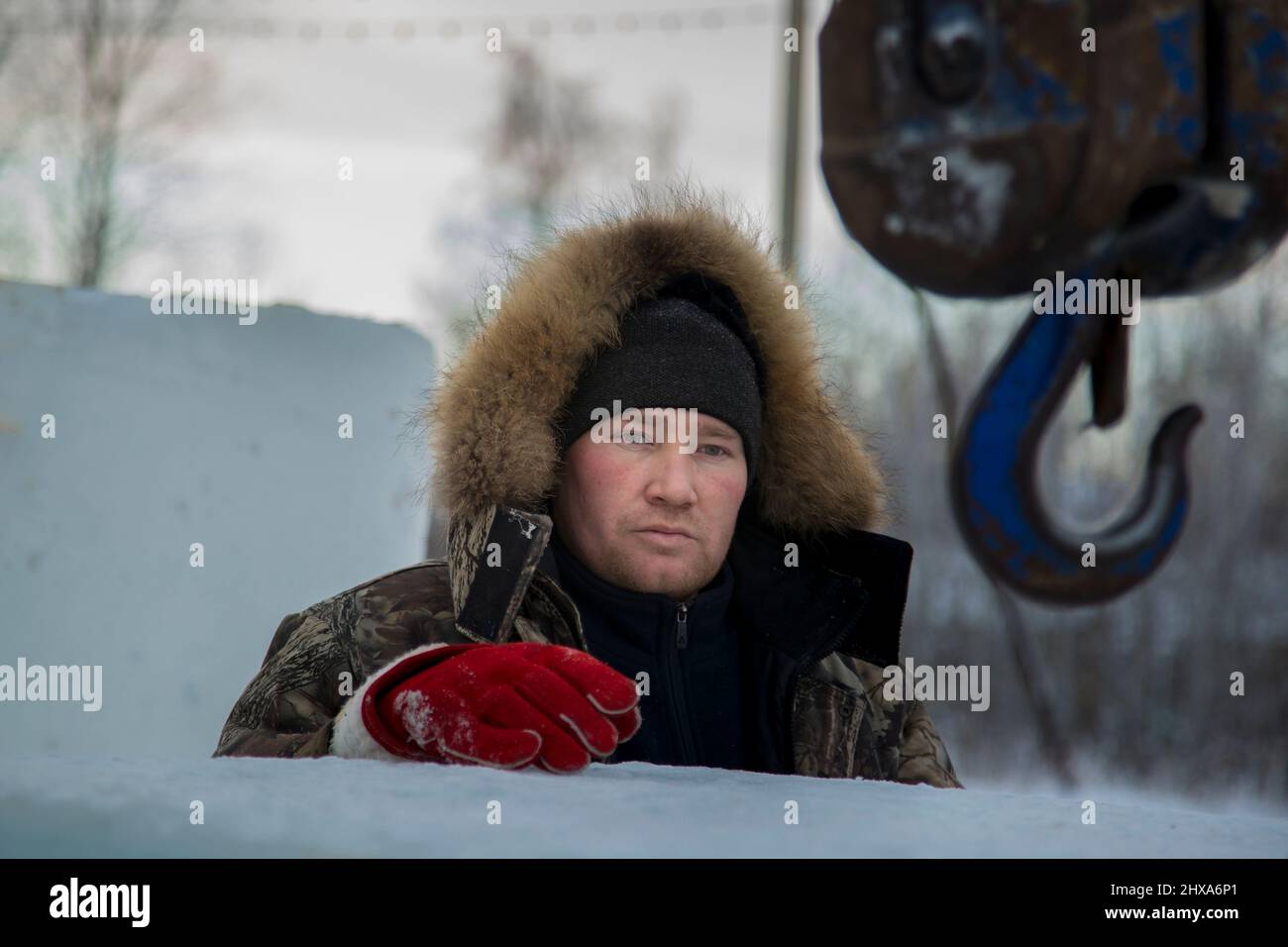 Porträt eines Arbeiters in einer Winterjacke mit einer Kapuze am Haken eines LKW-Krans beim Bau einer Eisstadt Stockfoto