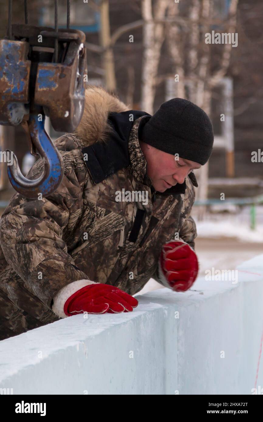 Porträt eines Arbeiters in einer Winterjacke mit einer Kapuze am Haken eines LKW-Krans beim Bau einer Eisstadt Stockfoto