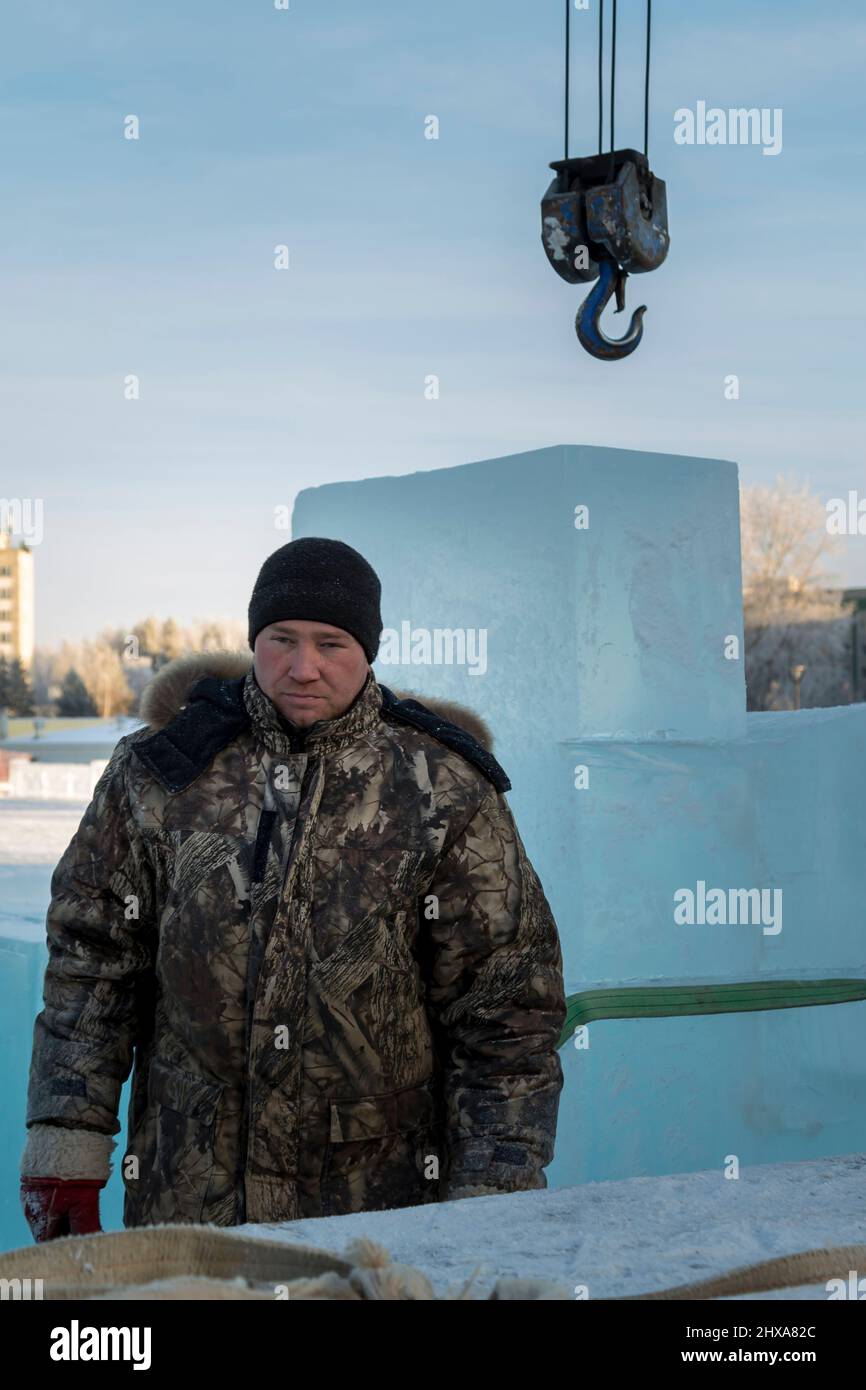 Porträt eines Arbeiters in einer Winterjacke mit einer Kapuze am Haken eines LKW-Krans, der Eisblöcke lädt Stockfoto