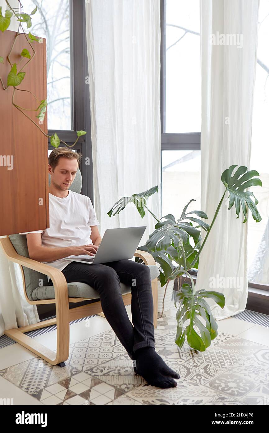 Surfen im Internet zu Hause. Hübscher junger Mann, der am Laptop arbeitet, während er zu Hause in einem großen bequemen Stuhl sitzt Stockfoto
