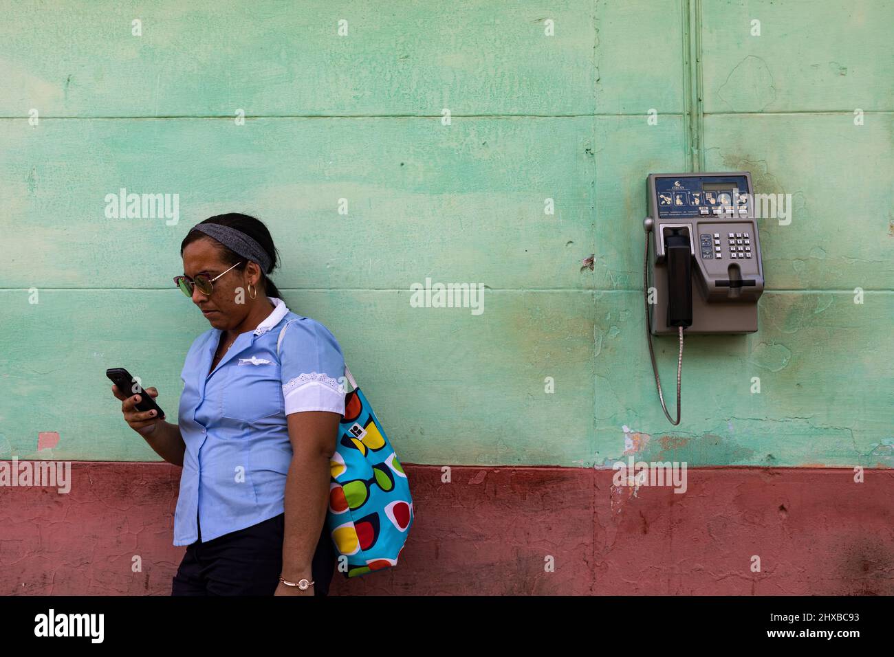 Eine Frau überprüft ihr Mobiltelefon, auf ihrem Rücken ein altes Telefon an der Wand Stockfoto