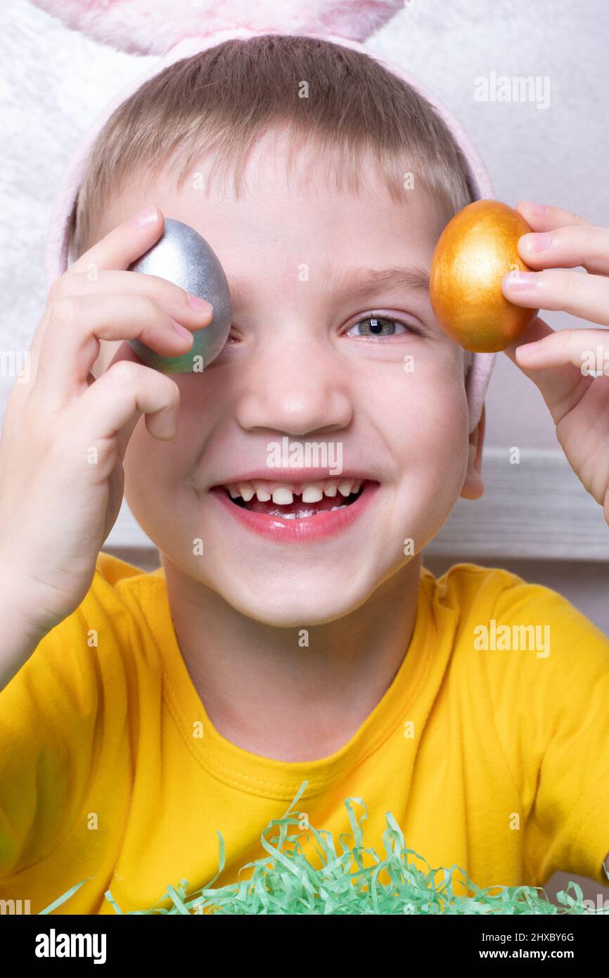 Porträt eines kleinen blonden Jungen, der seine Augen mit bunten Eiern bedeckt, Nahaufnahme, vertikaler Rahmen. Osterurlaub mit der Familie. Das Kind hat Spaß und feiert Stockfoto