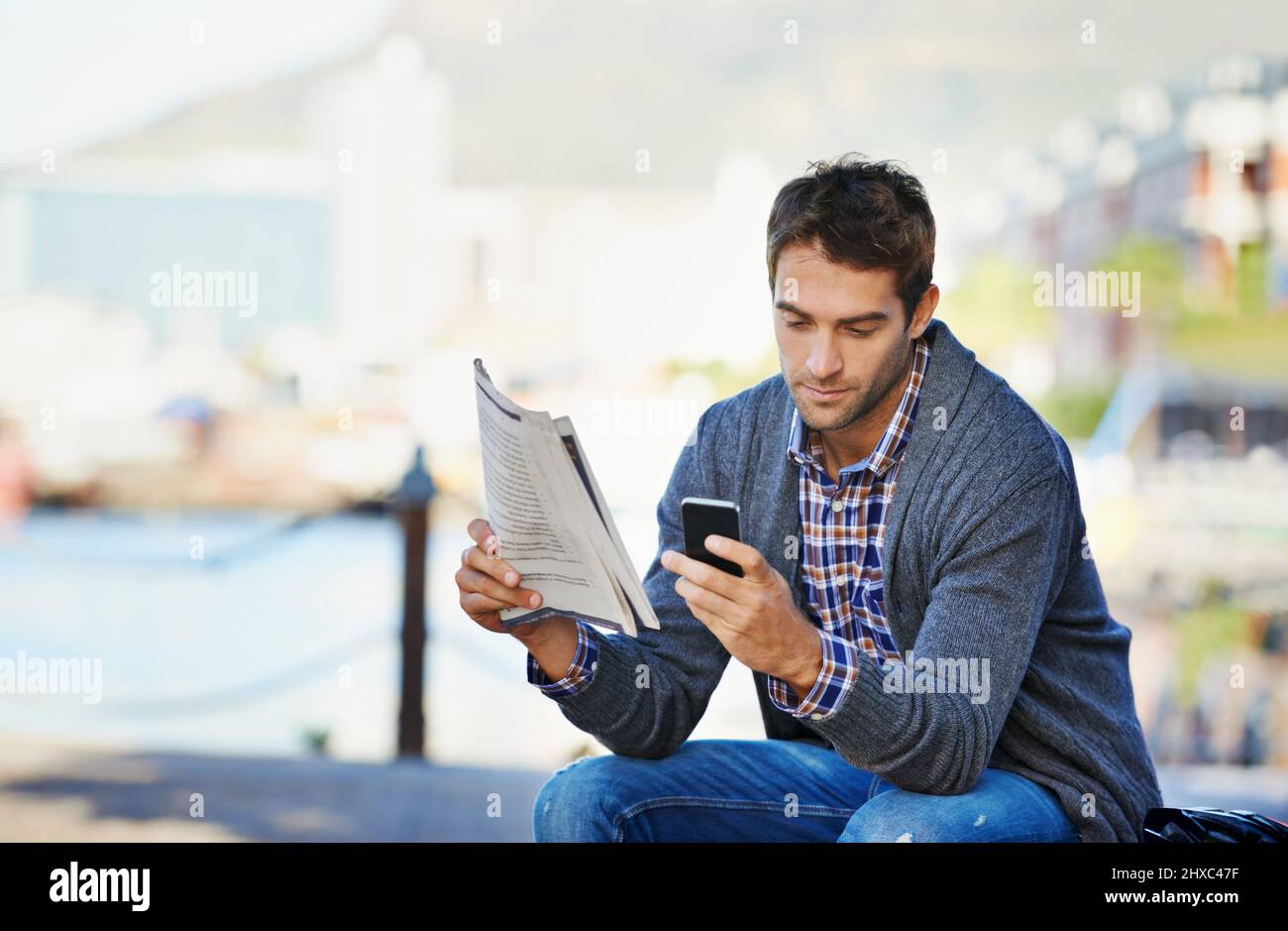 Neuposten der heutigen Nachrichten. Aufnahme eines hübschen Mannes, der die Zeitung hält und sein Telefon im Freien benutzt. Stockfoto