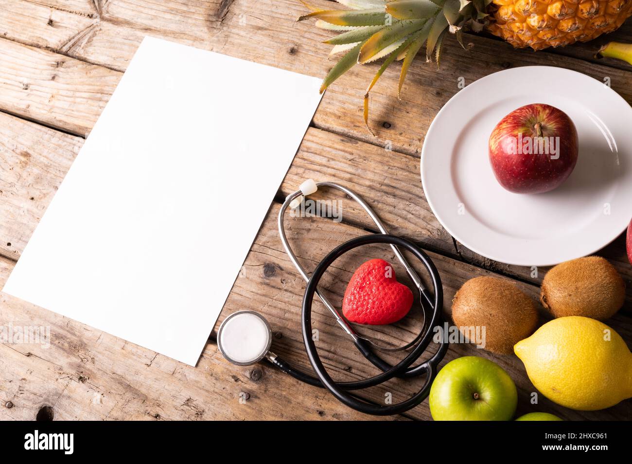 Hochwinkelansicht verschiedener Früchte mit Stethoskop und Herzform durch leeres Papier auf Holztisch Stockfoto