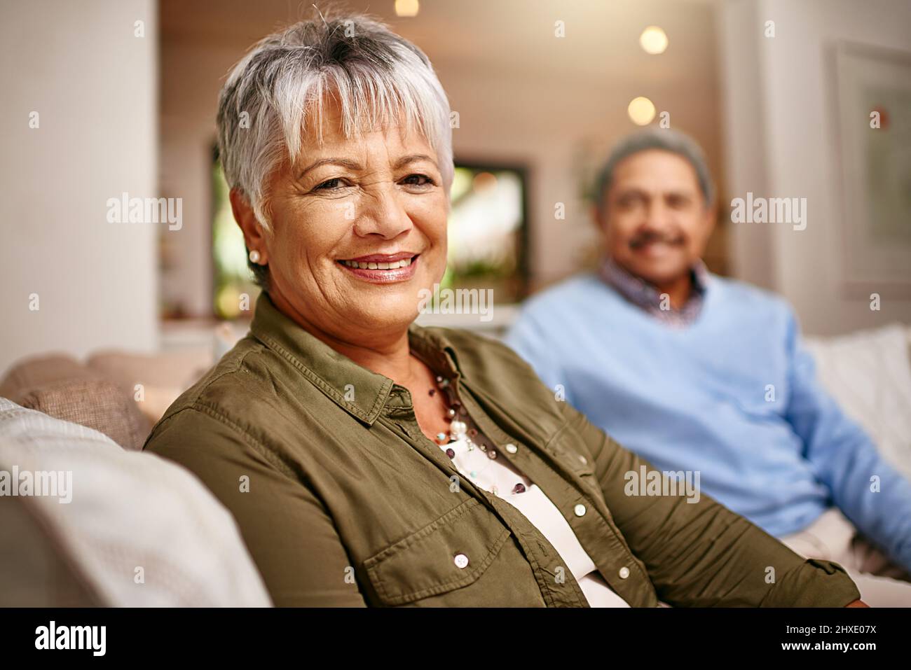 Wir haben es gut gemacht. Porträt einer glücklichen älteren Frau, die sich auf dem Sofa mit ihrem Mann im Hintergrund entspannt. Stockfoto