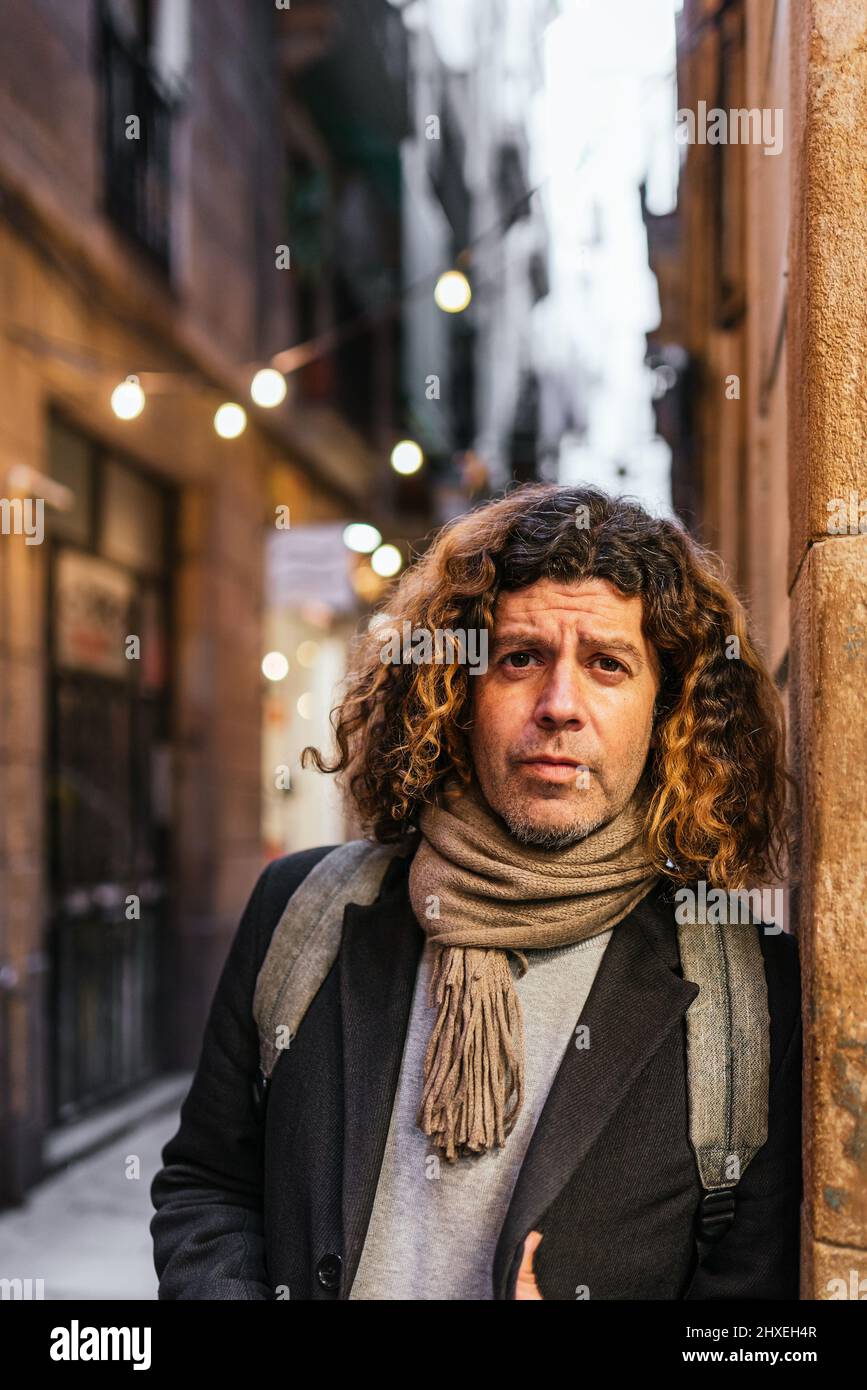 Ernsthafter Mann mittleren Alters in eleganter Freizeitkleidung, mit Rucksack und Schal, der in der Dämmerung auf der Stadtstraße auf die Kamera blickt und sich an der Wand lehnt Stockfoto