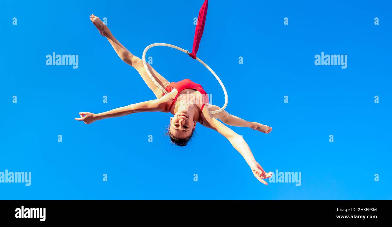 acrobat athletische, junge anmutige Turnerin Durchführung Luftübungen im Luftring im Freien auf Himmel Hintergrund. Flexible Frau in rotem Anzug führt Stockfoto