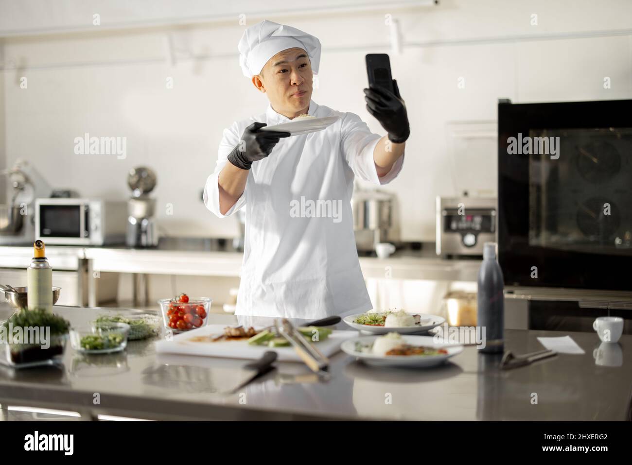 Der asiatische Koch zeichnete am Telefon einen kulinarischen Vlog auf und zeigte einen Teller mit gekochtem Essen in der Küche. Konzept von Video-Blogging und Kochen von Mahlzeiten für das Restaurant Stockfoto