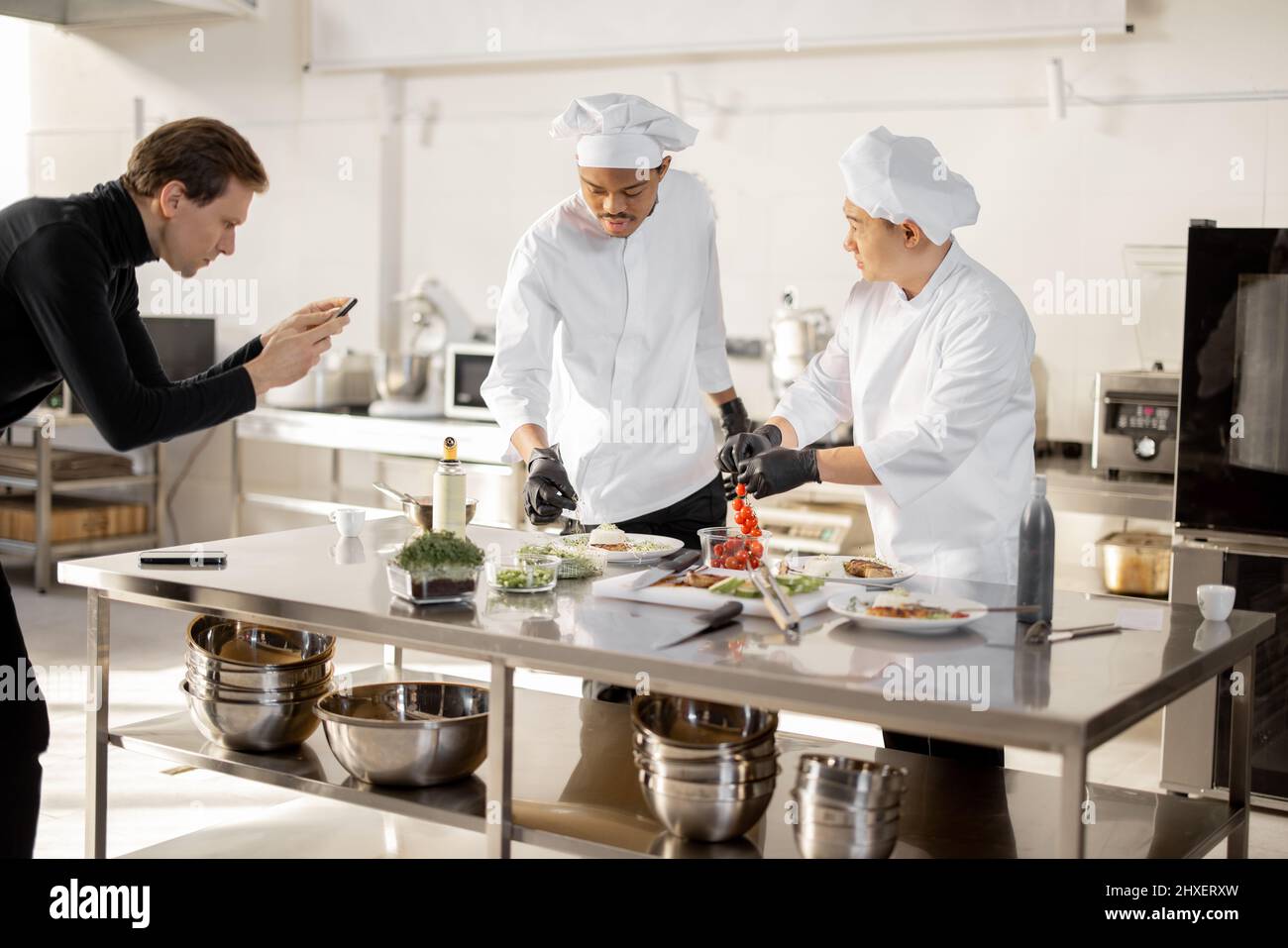 Mann zeichnet am Telefon den Prozess des Kochens von zwei Köchen in einer professionellen Küche auf. Lateinamerikanische und asiatische Köche bereiten Speisen für soziale Medien zu. Konzept des kulinarischen Videobloggens Stockfoto