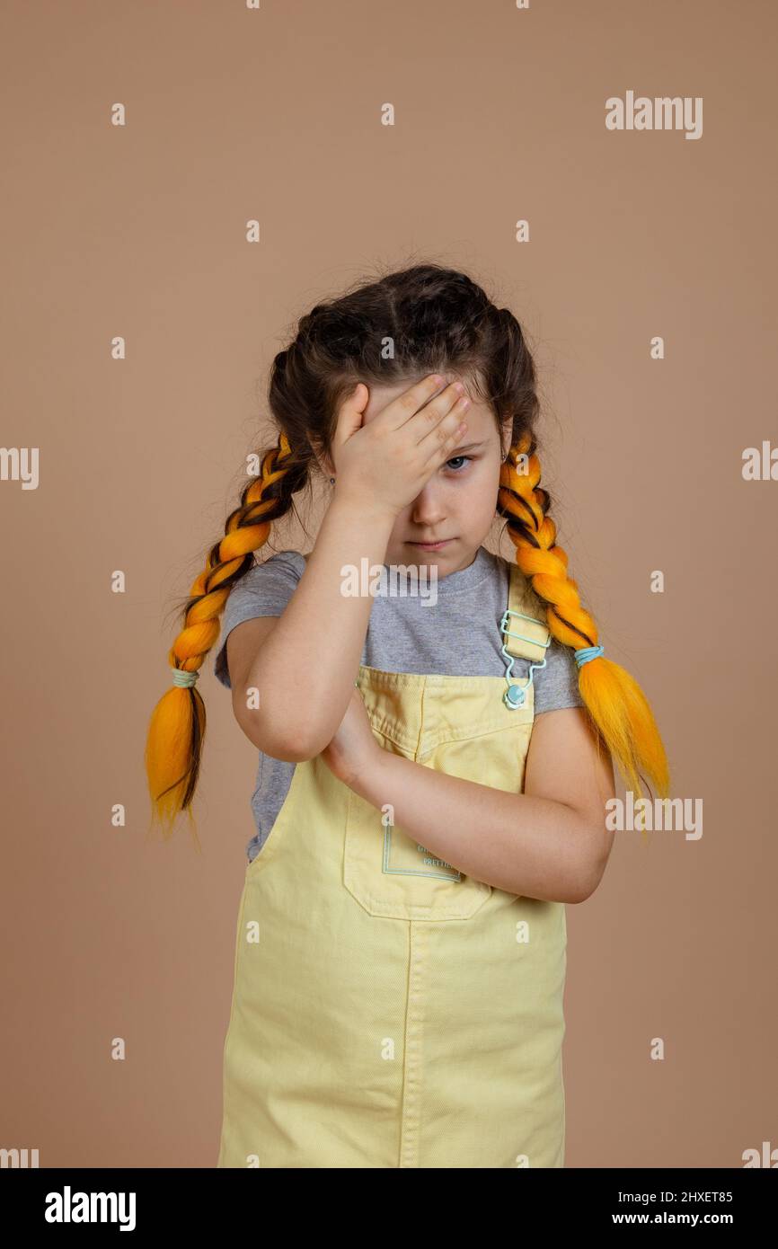 Kleines weibliches Kind mit gelben Kanekalon-Zöpfen schämt sich und berührt die Stirn mit der Hand, die ein Auge mit gelbem Jumpsuit und grauem T bedeckt Stockfoto