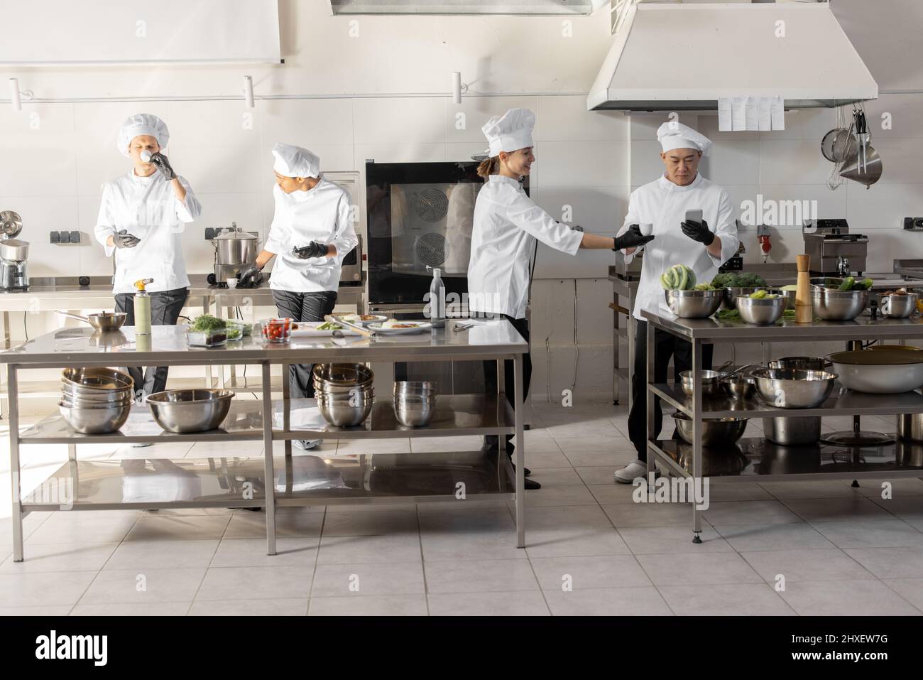 Gruppe von Köchen, die Telefone benutzen und während einer Pause in der Restaurantküche Kaffee trinken. Professionelles Personal ruht sich nach der Arbeit in der Küche aus. Ein Team von Köchen aus verschiedenen Rassen in Innenräumen Stockfoto