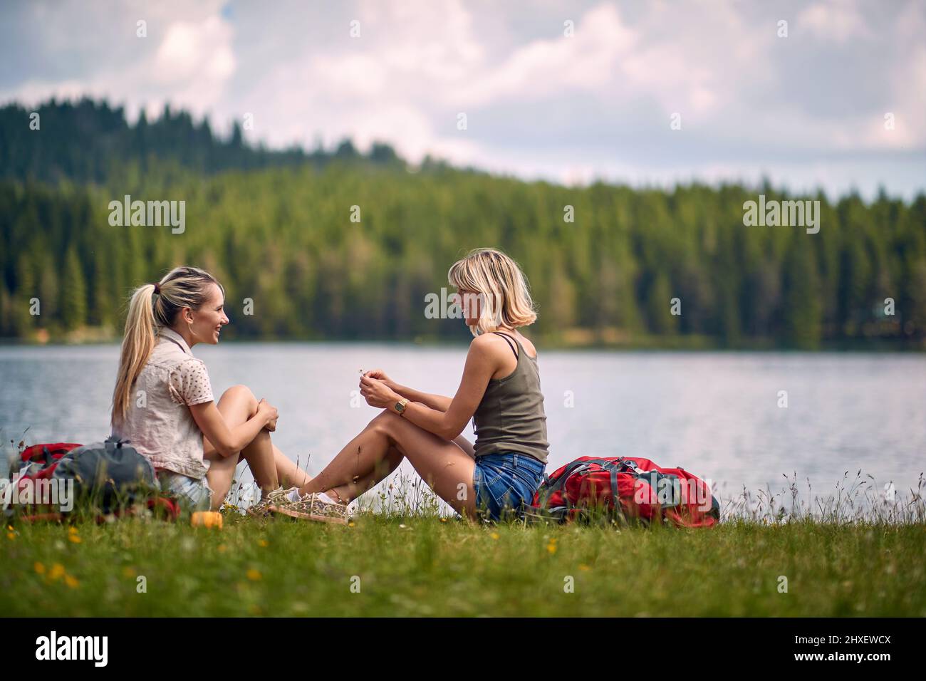 Zwei Frauen sitzen im Gras vor dem See und reden. Leirsure Zeit. Ausflug, Wochenende, Spaß, Lifestyle, Naturkonzept Stockfoto