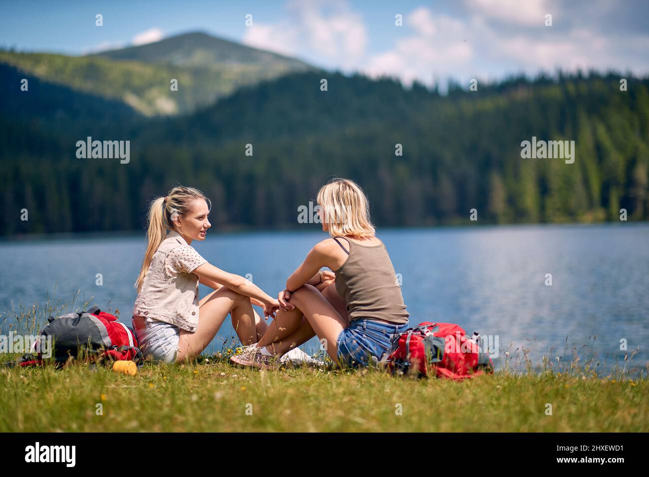Zwei junge Freundinnen sitzen im Gras vor dem See und reden miteinander, Spaß, Lifestyle, Naturkonzept Stockfoto
