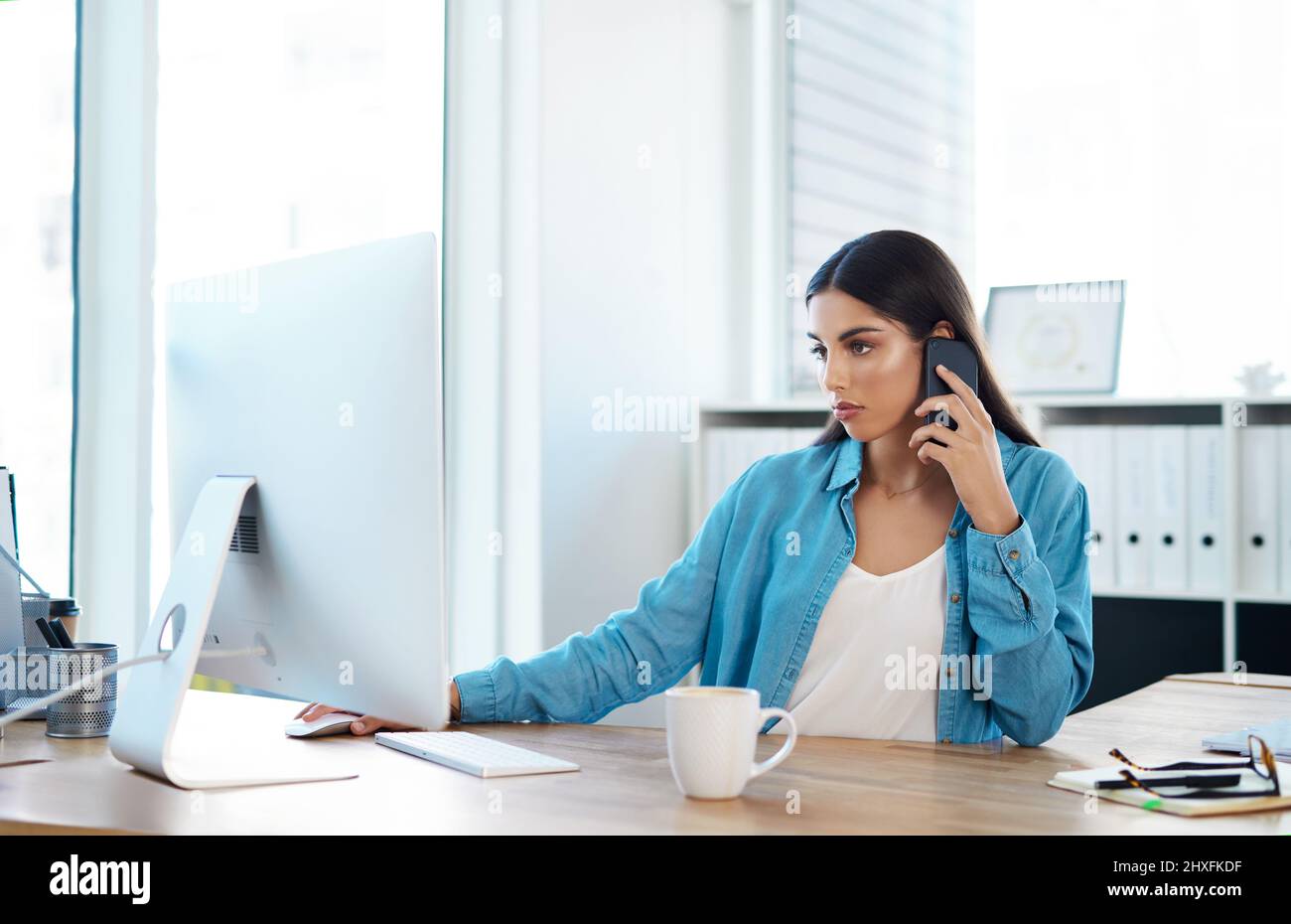 Management eines kleinen, aber schnell wachsenden Unternehmens. Aufnahme einer jungen Geschäftsfrau, die auf einem Mobiltelefon spricht, während sie an einem Computer in einem Büro arbeitet. Stockfoto