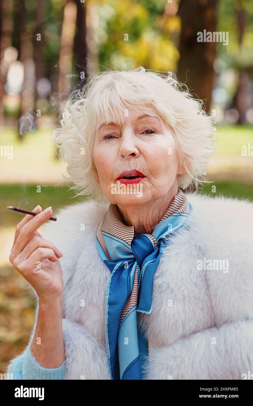 Lächelnd Senior elegant stilvolle modische Frau mit grauen Haaren in Pelzmantel im Freien Rauchen Zigarette. Ungesunder Lebensstil, Alter, positive Vibes Stockfoto