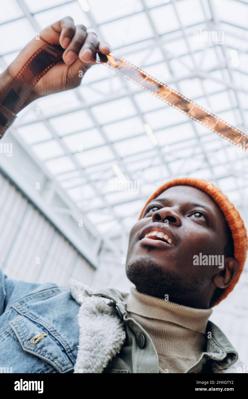 Emotional glücklicher afroamerikanischer Männerfotograf in Denim-Jacke blickt in hell erleuchteter, extremer Nahaufnahme auf Vintage-Film Stockfoto