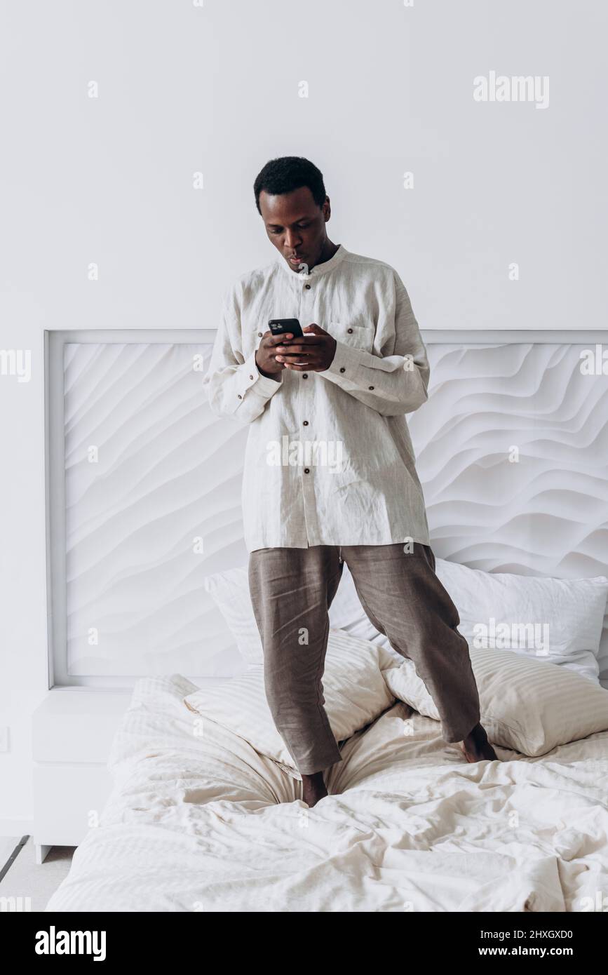 Ein lustiger, aufgeregter junger afroamerikanischer Mann in legerer Kleidung springt in einem modernen Zimmer mit Mobiltelefon auf einem großen Bett hoch Stockfoto