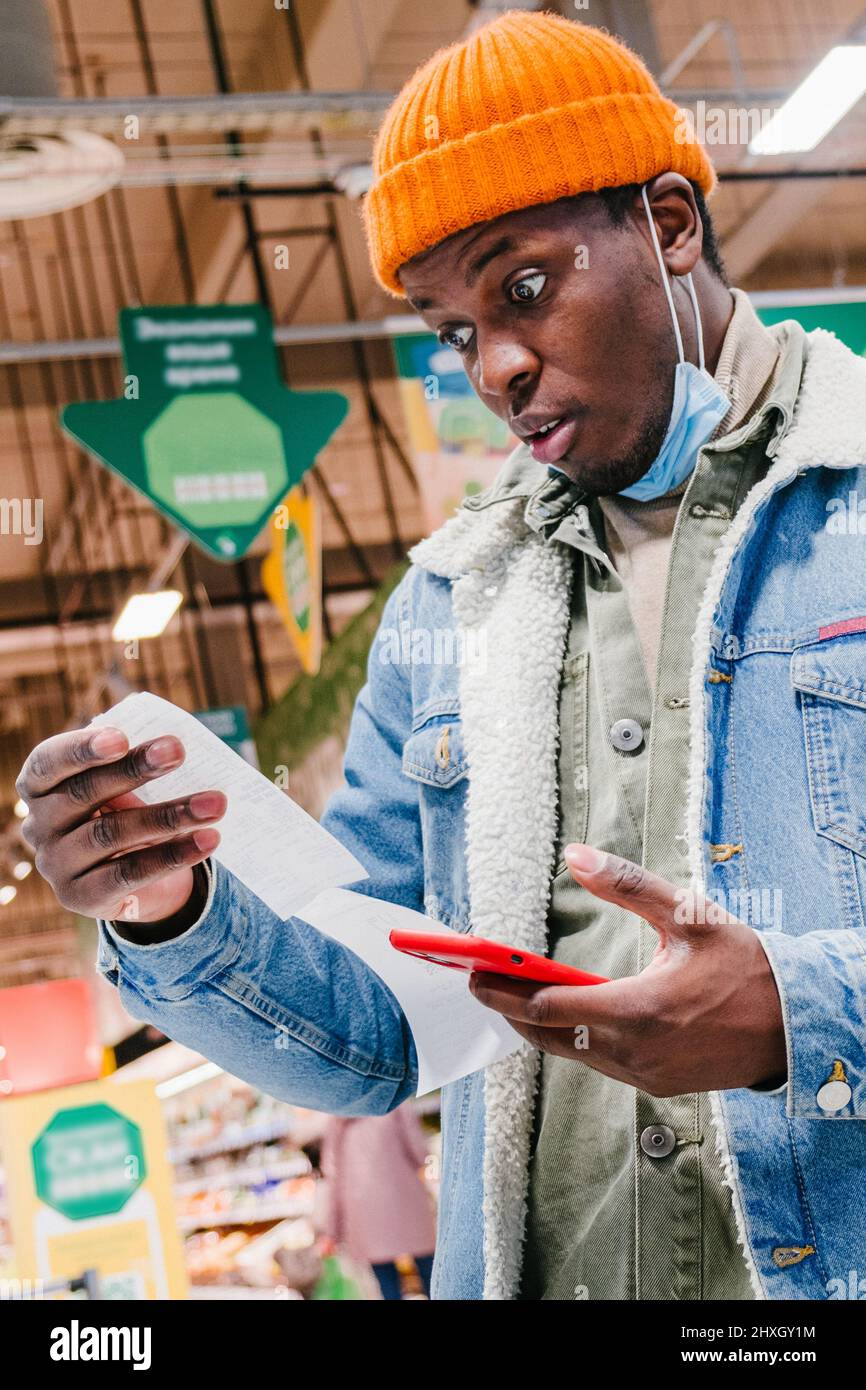 Enttäuschter afroamerikanischer Kunde in Denim-Jacke mit Maske sieht auf Quittung total Mobiltelefon im Supermarkt halten Stockfoto