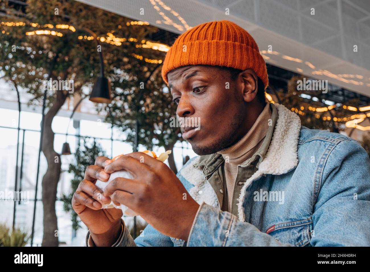 Ein netter afroamerikanischer Mann in orangefarbenem Strickhut isst in einem modern eingerichteten Café aus der Nähe einen köstlichen frischen Hamburger Stockfoto