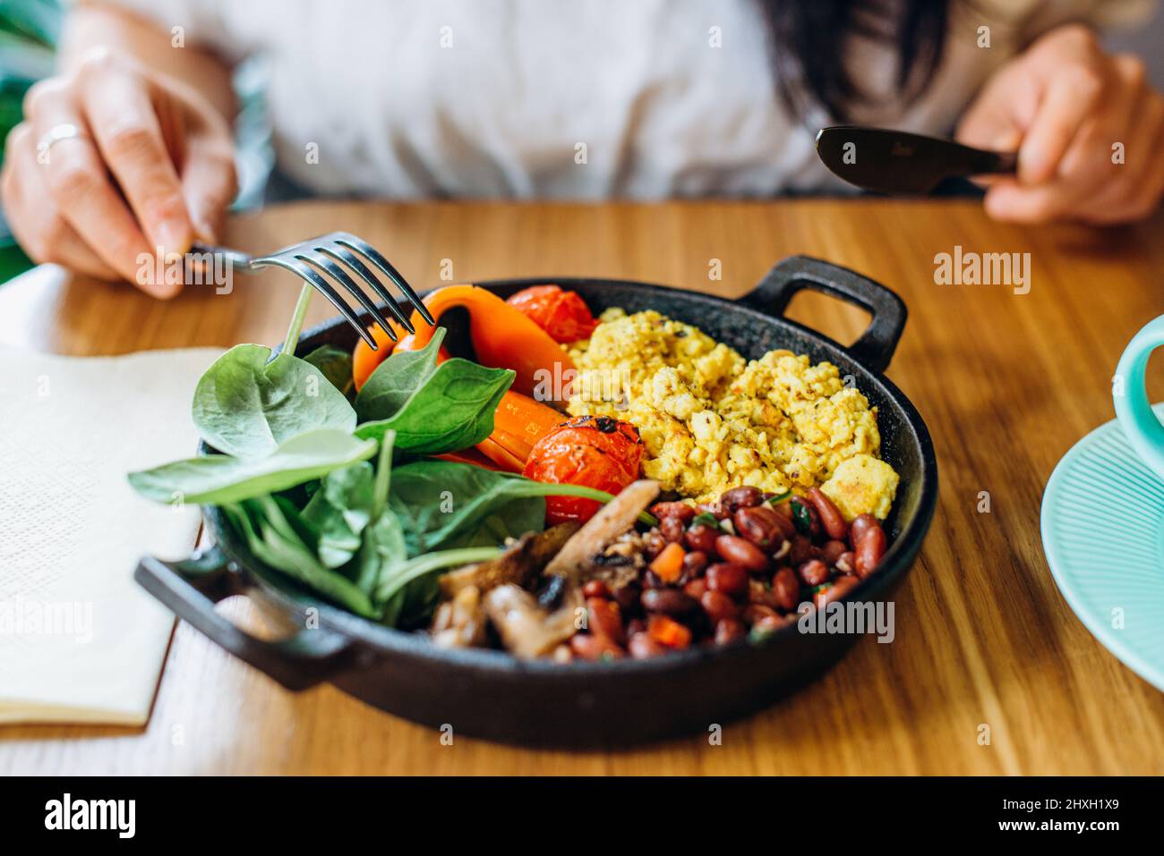 Veganes Gericht aus verschiedenen Gemüsesorten auf der Bratpfanne und Frauenhände mit Gabel und Messer am Tisch im Cafe aus der Nähe Stockfoto