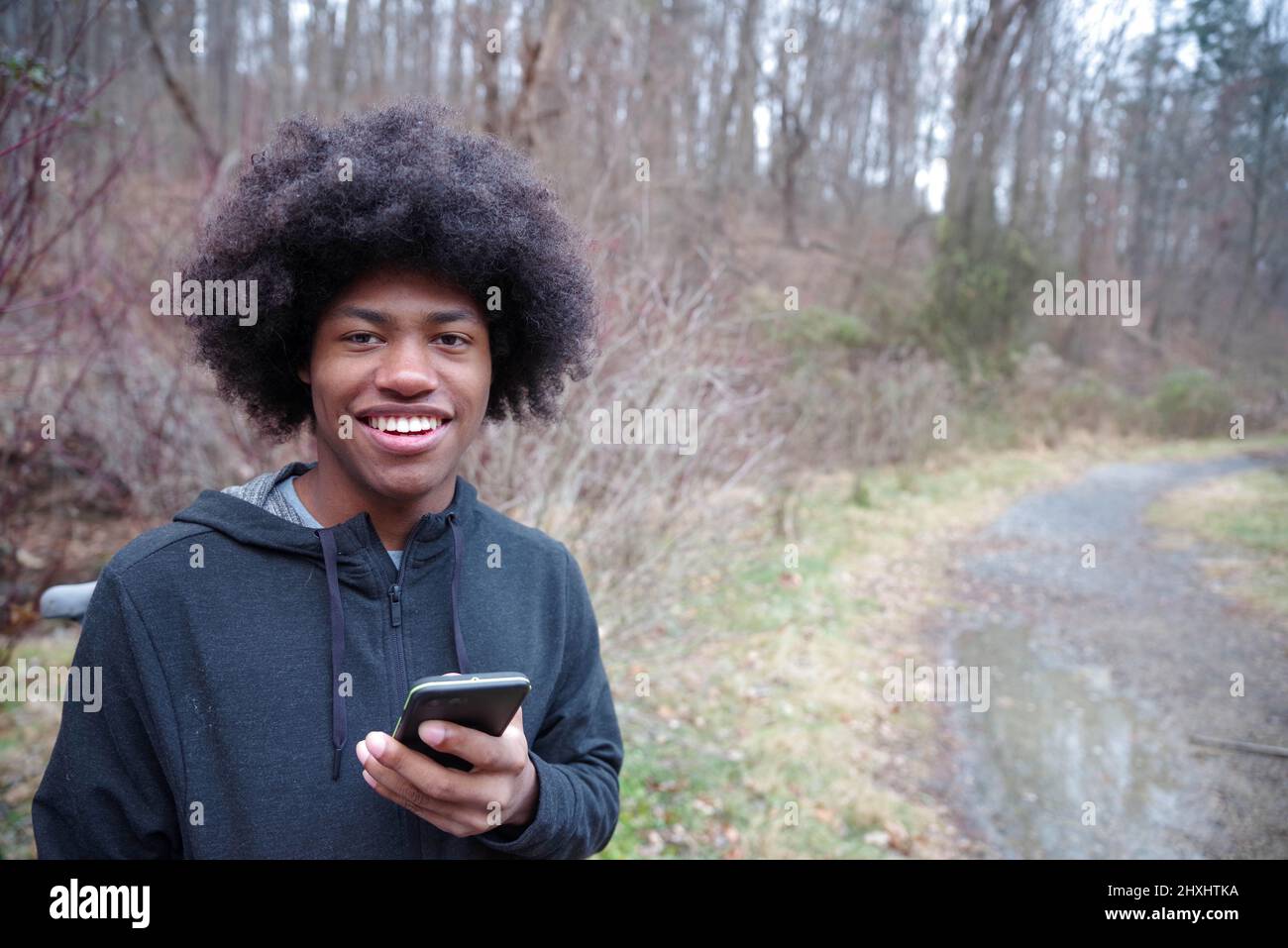 Afroamerikanischer Teenager lächelt mit seinem Telefon auf einem bewaldeten Wanderweg Stockfoto