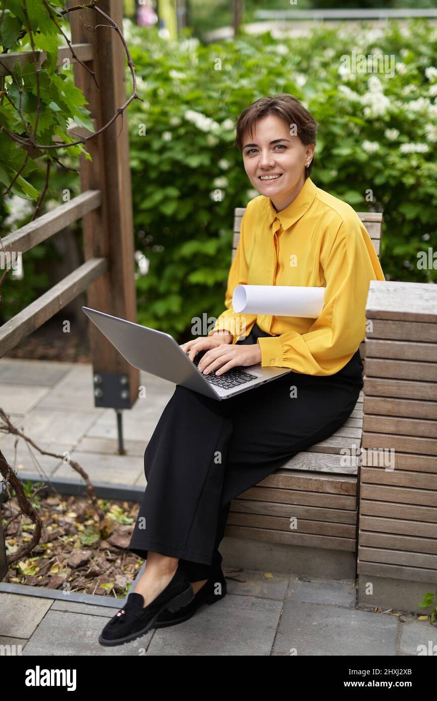 Business, Finanzindustrie oder Immobilienkonzept. Lächelnde, erfolgreiche Geschäftsfrau in gelber Bluse, die auf einer Bank im Freien im Park sitzt und einen Laptop benutzt, der bei der Arbeit auf die Kamera schaut. Hochwertige Bilder Stockfoto