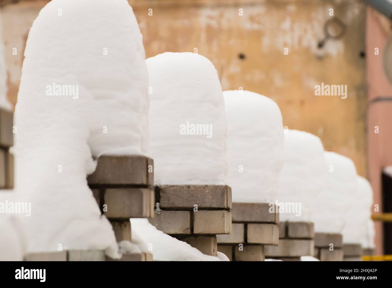 Große, schöne Säulen aus weißem Schnee auf Ziegelsteinsäulen. Eine gerade Reihe kleiner Schneeverwehungen vor dem Hintergrund einer schäbigen Wand. Urbane Winterlandschaft. Wolkiger Wintertag, weiches Licht. Stockfoto