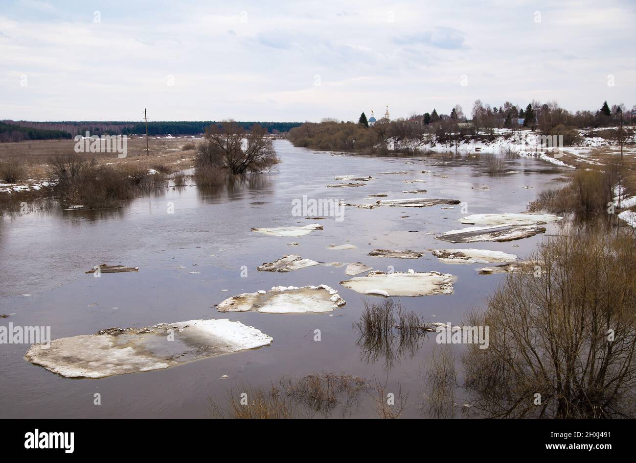 Kleine weiße Eisschollen Reihen sich in einer Linie aneinander und schweben den Fluss hinunter. Frühling, Schnee schmilzt, trockenes Gras rundherum, Überschwemmungen beginnen und der Fluss überfließt. Tag, bewölktes Wetter, weiches warmes Licht. Stockfoto