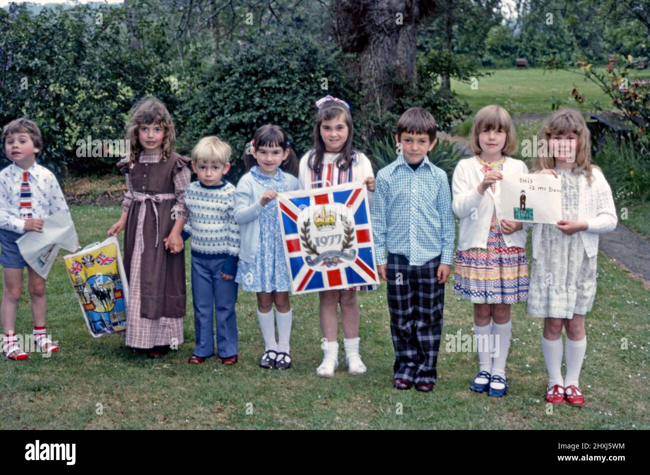 Eine Gruppe britischer Kinder, die am 7. Juni 1977 zusammenkommen, um das Silberjubiläum von Königin Elizabeth II. Zu feiern Hier halten die Kinder Plastikträger mit Jubilee-Bildern auf sich. Seltsamerweise halten zwei Kinder eine Zeichnung der TV-Comedy-Figur Mr Bean (die Worte lauten ‘This is ‘Mr Bene’). Dieses Bild stammt von einem alten Kodak-Amateurfotograf mit Farbtransparenz – einem Vintage-Foto aus dem Jahr 1970s. Stockfoto