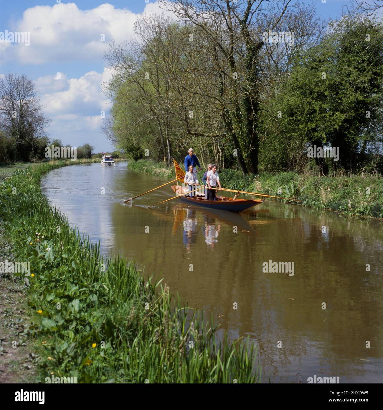 Vier Personen rudern ein Boot mit großer Flagge am Heck, während sie auf dem Kennet & Avon Kanal in der Nähe von All Cannings, Wiltshire, England, stehen Stockfoto