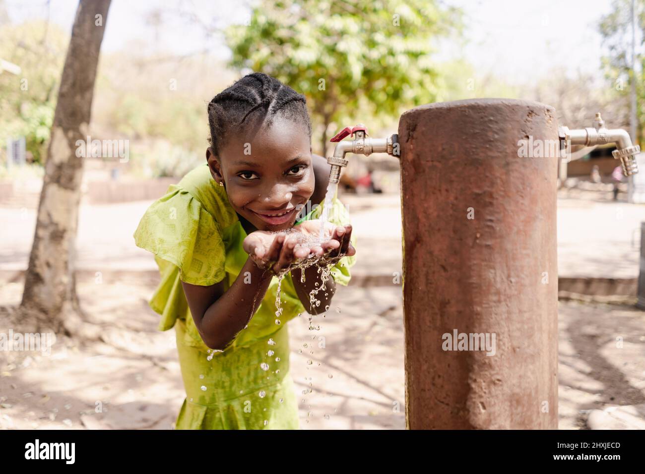 Fröhliches schwarzes afrikanisches Mädchen in einem schönen gelben Kleid, das frisches Wasser aus einem öffentlichen Wasserhahn trinkt Stockfoto