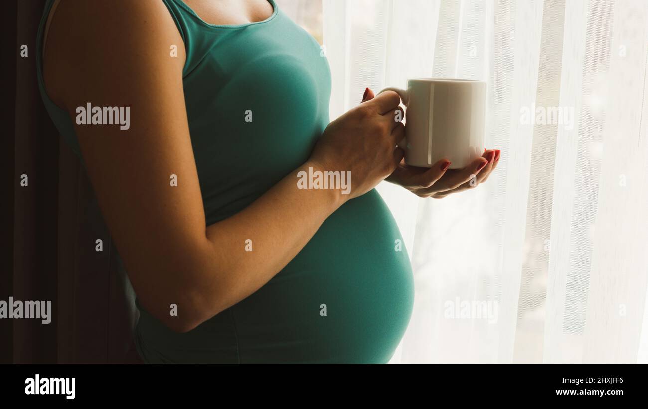 Mug Mockup. Schwangere mit weißem Becher. Schwangere trinkt Kaffee am Fenster. 6 Monate schwanger Frau trinken heißen Tee stehend Stockfoto
