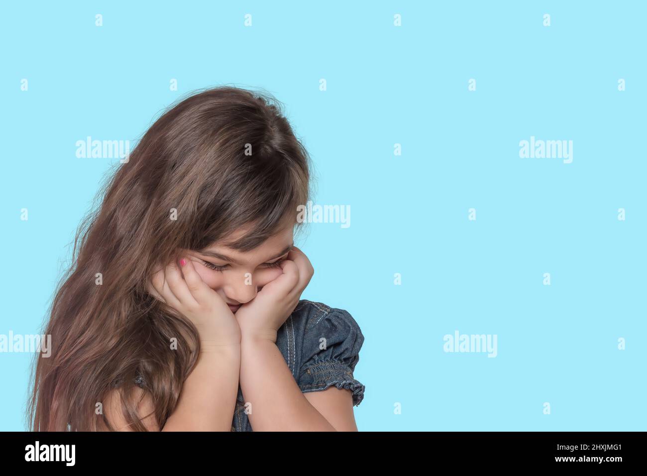 Beschämt gebräunte langhaarige kleine Mädchen trägt Denim-Kostüm posiert auf cyanfarbenem Hintergrund. Stockfoto