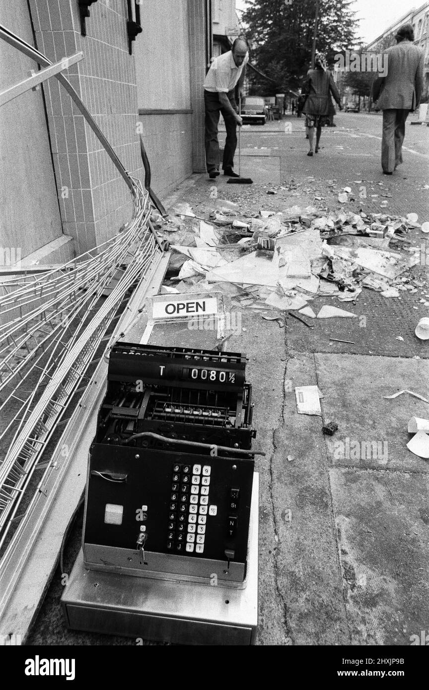 Die Säuberung begann in den Straßen von Notting Hill nach den Unruhen am montagabend nach dem Karneval. Ein Co-Op-Mitarbeiter beginnt die große Aufgabe, die Trümmer zu fegen, im Vordergrund eine zertrümmerte Kasse aus dem Geschäft. 31.. August 1976. Stockfoto