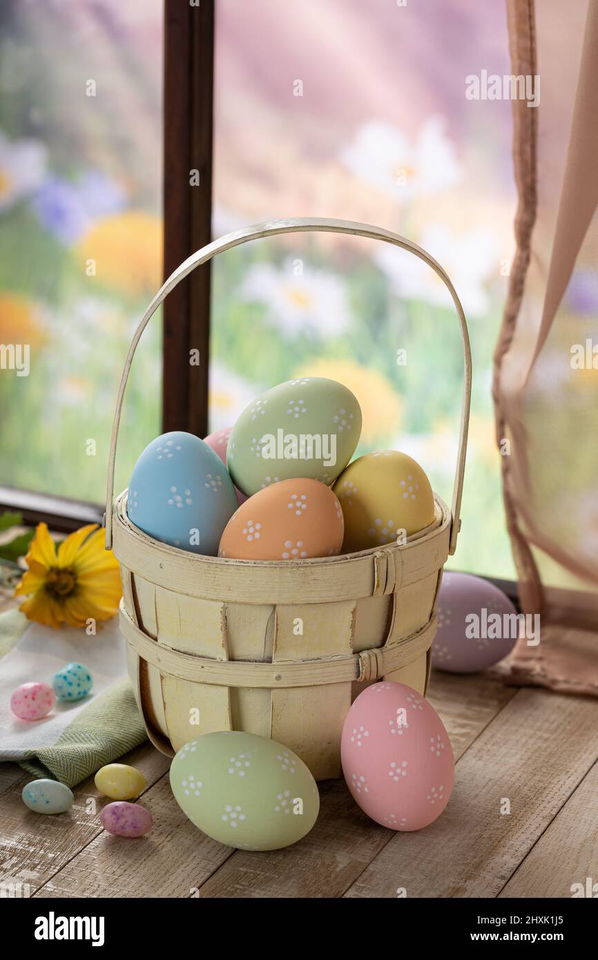 Bunte ostereier in einem Korb und auf einem Holztisch an einem Fenster mit buntem Frühlingshintergrund Stockfoto