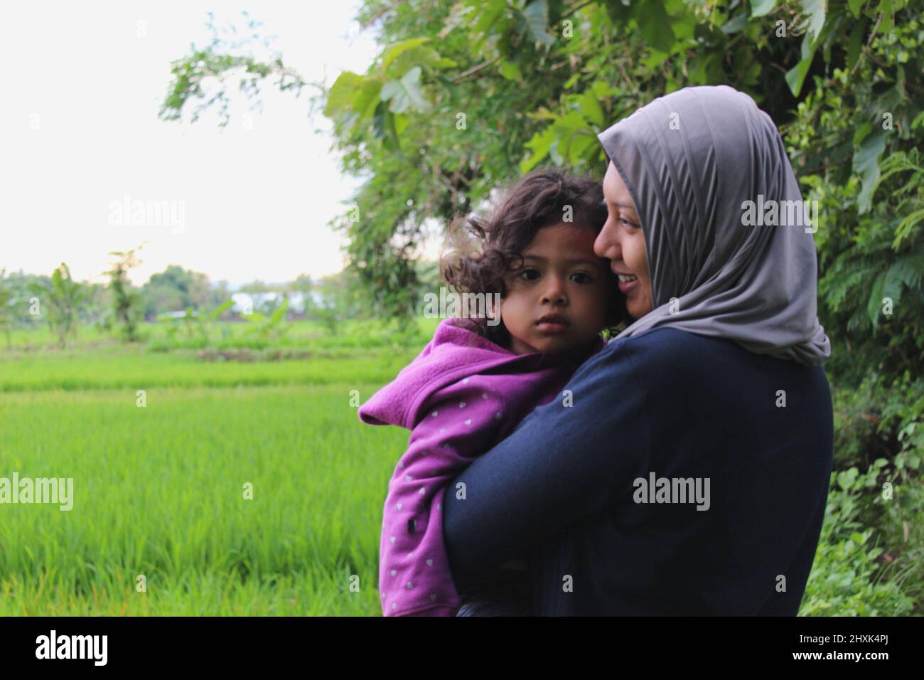 Java, Indonesien - 12 24 2020: Verschwommene Hintergrundansicht von Reisfeldern und einer Frau in einem Hijab, die ein Mädchen hält, sieht glücklich aus Stockfoto