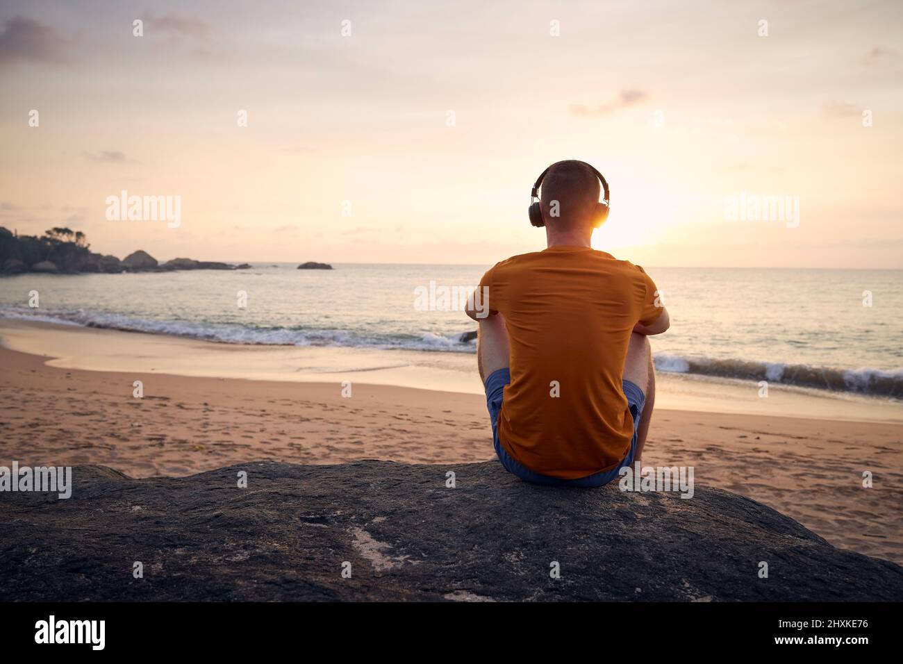 Rückansicht des Mannes mit Kopfhörern am Strand. Gelassenheit, Kontemplation und Musikhören bei schönem Sonnenuntergang. Stockfoto
