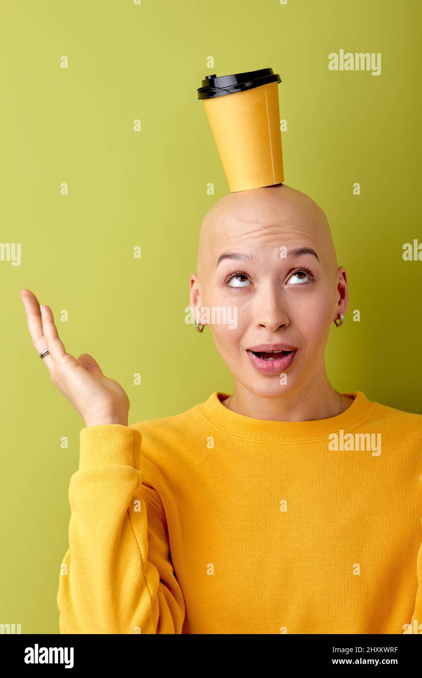 Wunderschöne Glatze kaukasische Frau mit Take-away-Kaffee-Tasse auf dem Kopf isoliert über grünem Hintergrund. Junge überrascht Frau in gelbem Hemd posiert vor der Kamera Stockfoto