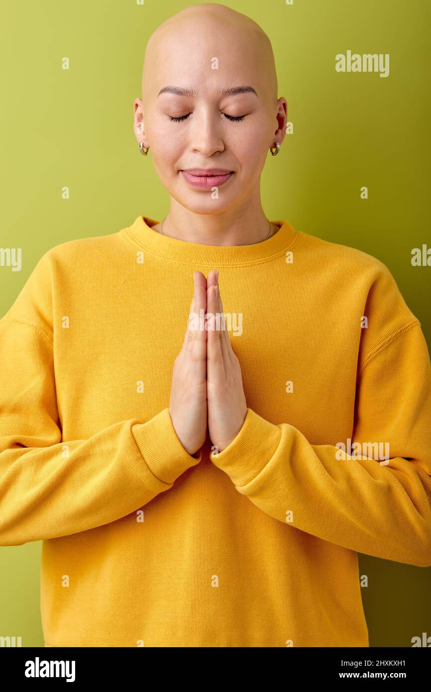 Glatze Dame faltet die Hände in Gebetsstellung. Isolierter grüner Studiohintergrund. Beten, lächeln. Porträt von kaukasischen schönen weiblichen Modell. Menschliche EM Stockfoto