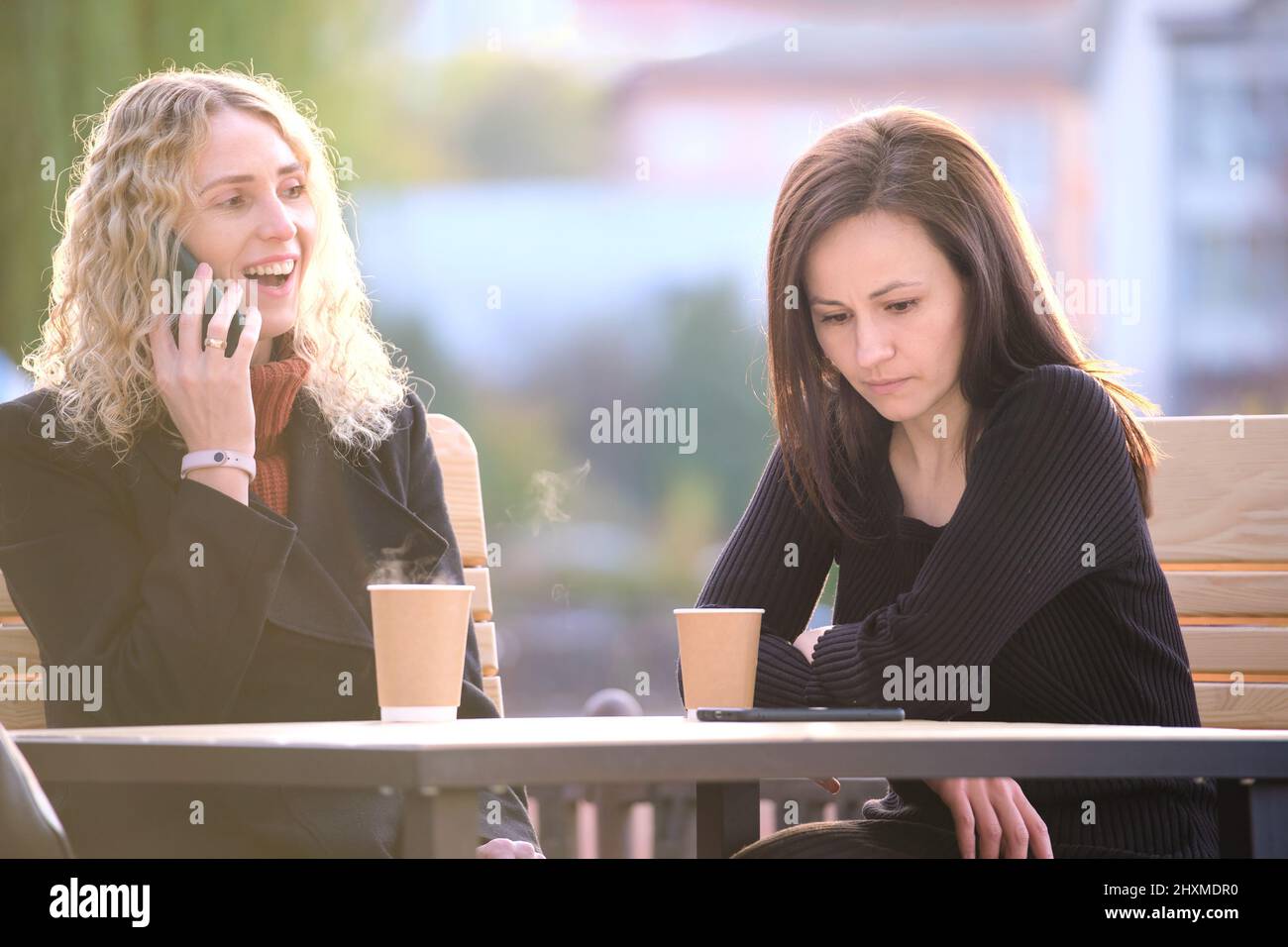 Traurige Frau, die von ihrer Freundin ignoriert wird, die im Straßencafé im Freien sitzt, während sie glücklich auf dem Handy redet und keine Aufmerksamkeit schenkt. Freundschaft Stockfoto
